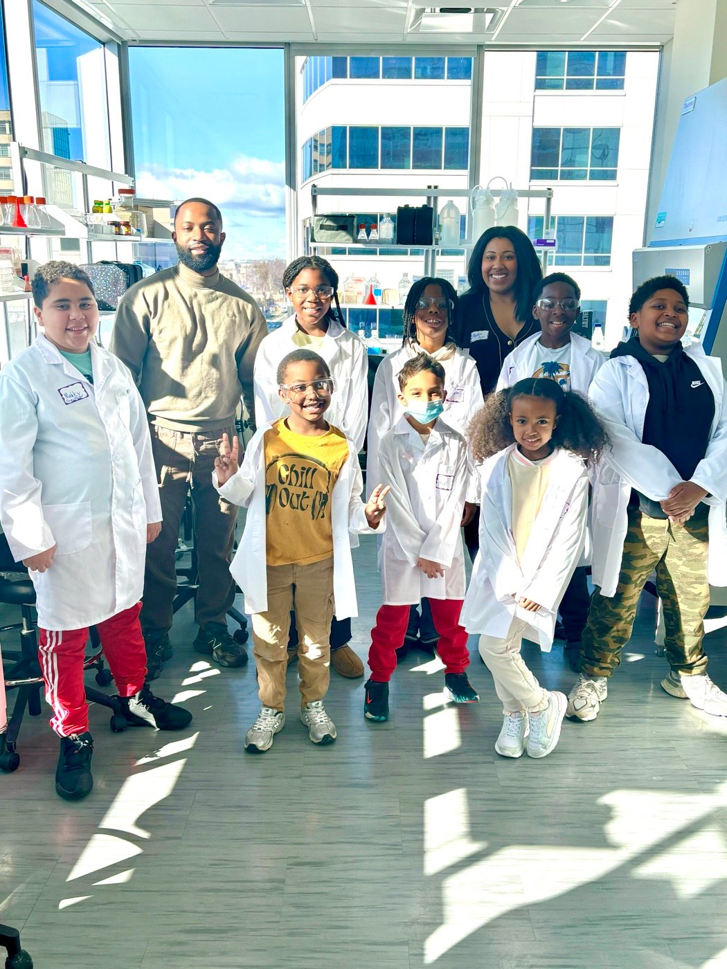 Group of young students in lab coats posing with Myettia in a lab